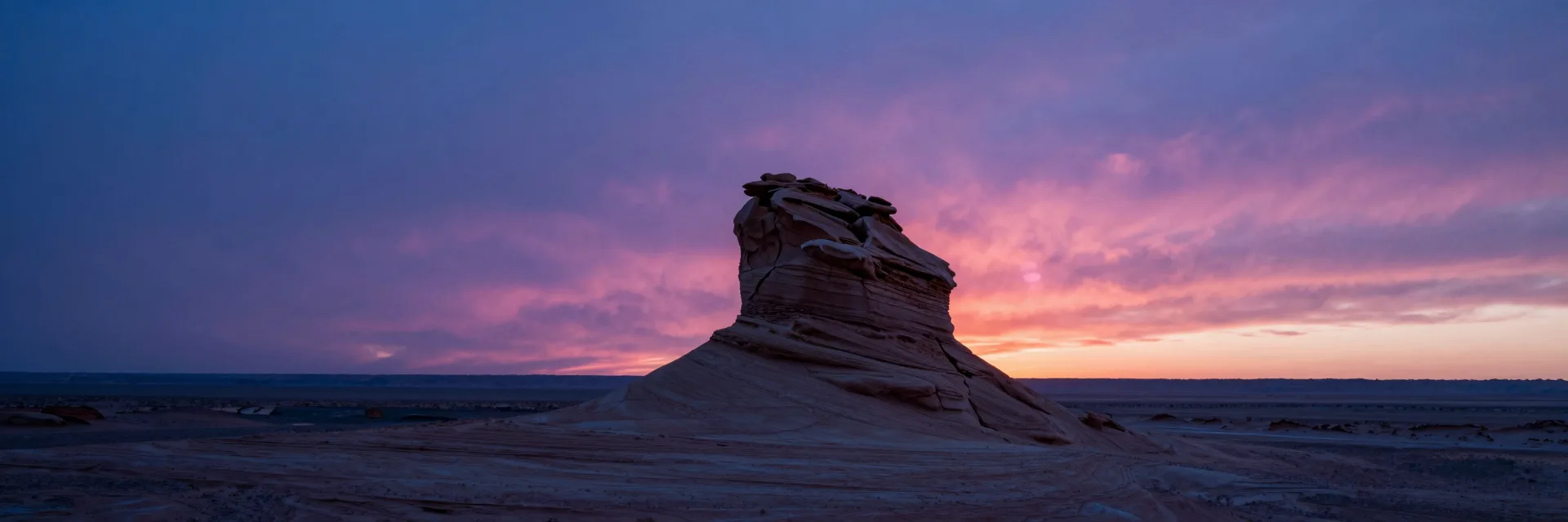 Single sandstone formation small in the right third, dwarfed by a vast pink and purple sunset sky filling the upper two-thirds, isolated against the desert horizon.