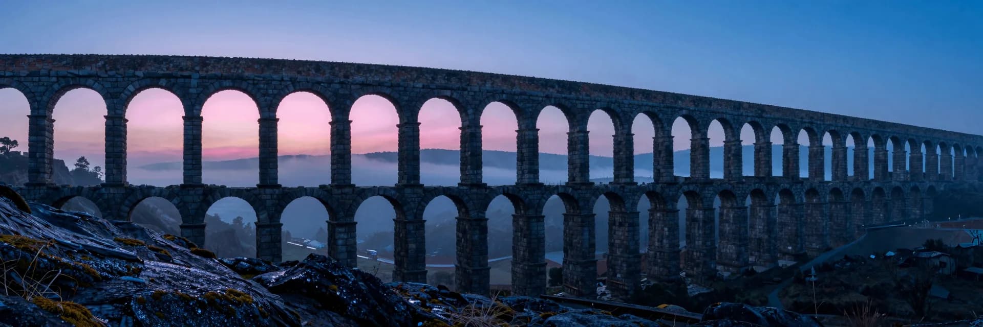Ultra-wide panorama of a multi-tier stone aqueduct receding into deep distance across a misty valley at dawn, arches rhythmically smaller as they recede, hot-pink dawn glow on the highest crests.