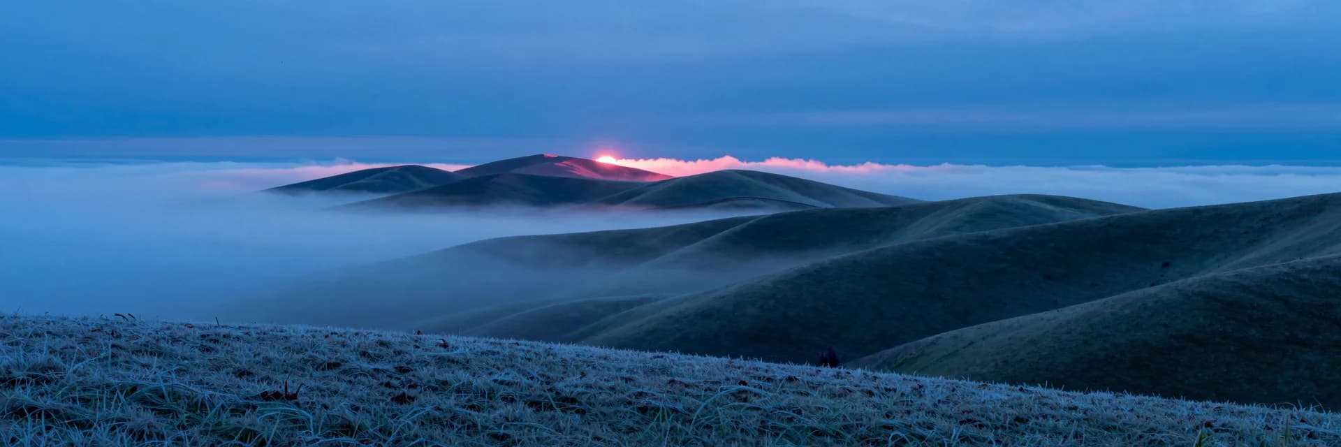 Ultra-wide panoramic landscape of rolling green-blue hills at dawn with a thick horizontal fog band obscuring the middle distance, hills emerging above and below.