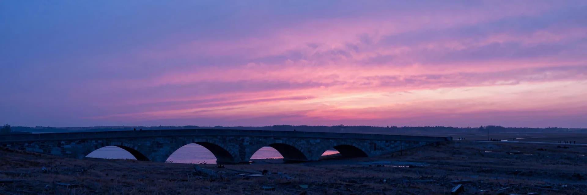 Ultra-wide distant view of the stone arch bridge as a small element in a vast landscape, dramatic horizontal scale.