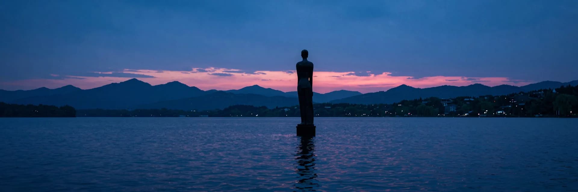 Ultra-wide composition of a small sculpture standing alone in a vast lake and mountain landscape.