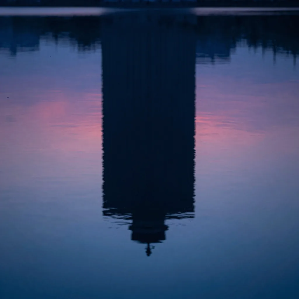 Still water surface mirroring the silhouette of a sculpture and the gradient of a twilight sky.