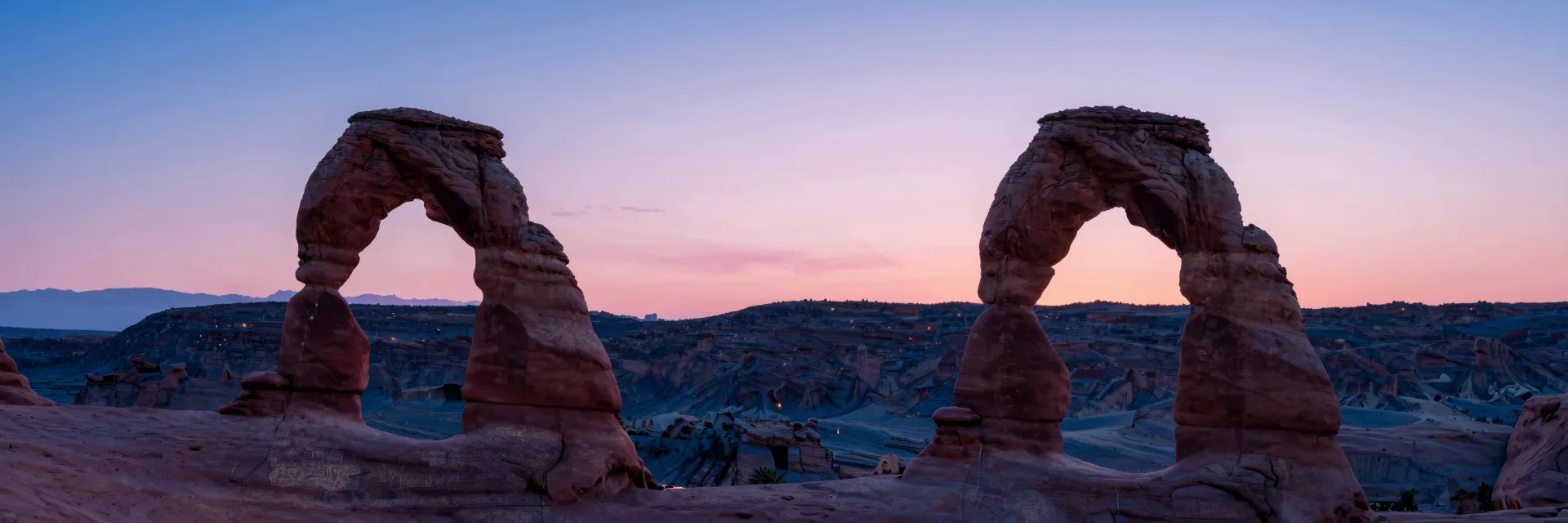 Ultra-wide vista of three small sandstone arches as distant elements in a vast desert landscape at twilight